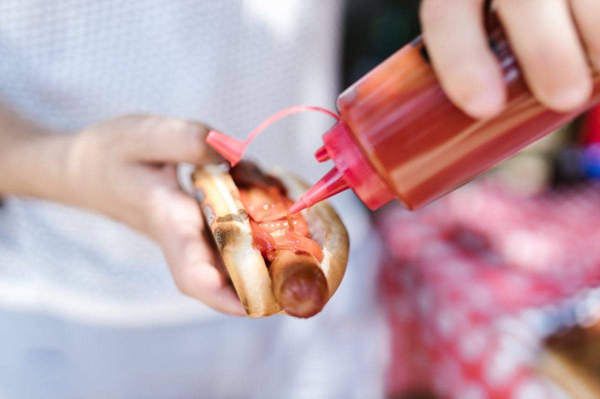 Seseorang menuangkan saus tomat di atas hot dog dengan roti panggang menggunakan botol saus berwarna merah.