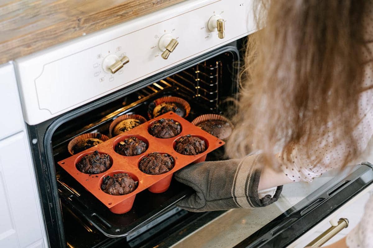 Seseorang mengenakan sarung tangan oven sedang mengeluarkan loyang berisi muffin cokelat dari oven dapur modern.