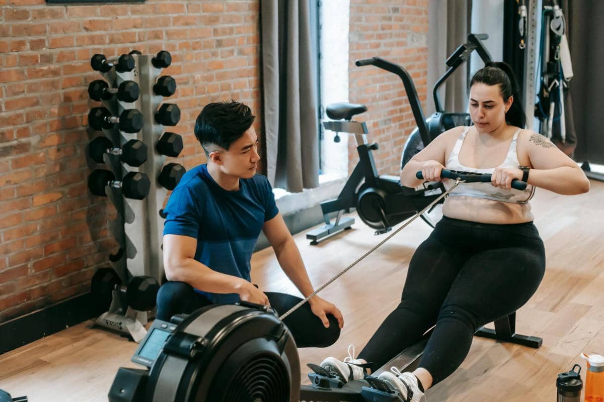 Seorang perempuan berolahraga dengan mesin dayung di gym sambil didampingi pelatih pria yang memberikan arahan latihan.