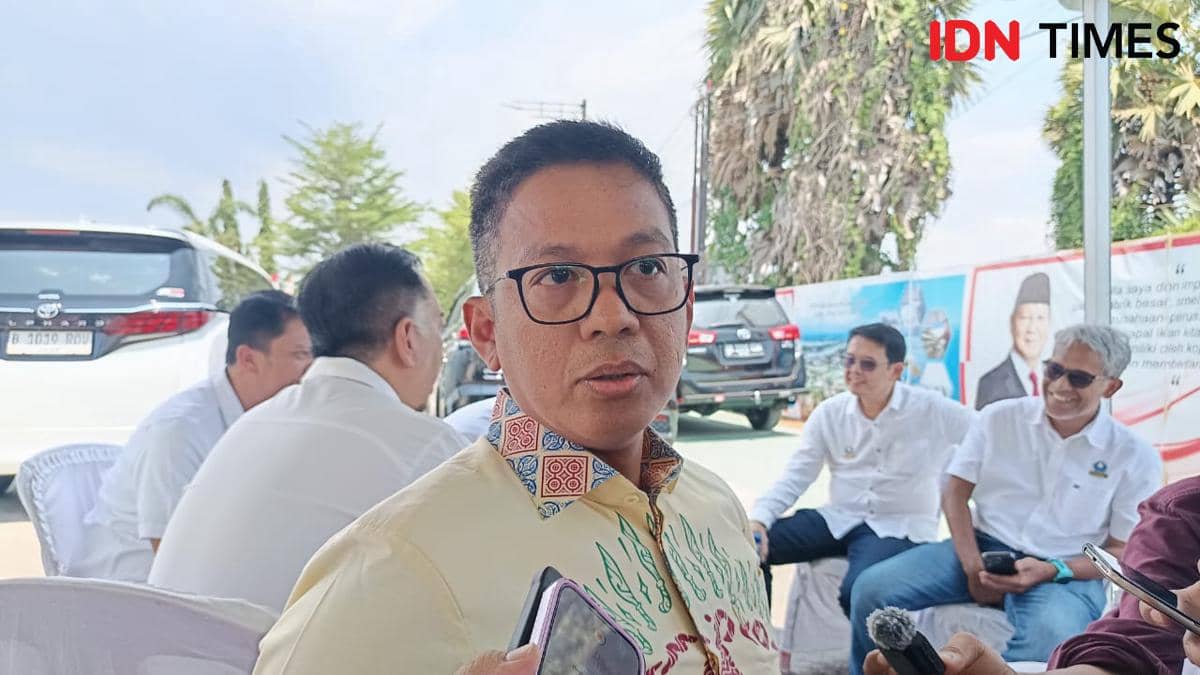 Seorang pria berkacamata mengenakan kemeja bermotif batik sedang berbicara di luar ruangan dengan beberapa orang di latar belakang.