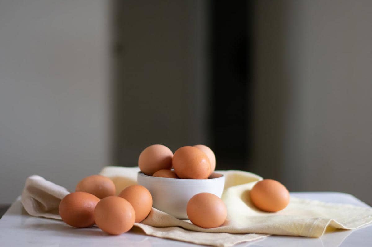 Beberapa butir telur berwarna cokelat diletakkan di dalam mangkuk putih dan di atas kain berwarna krem di meja dapur.