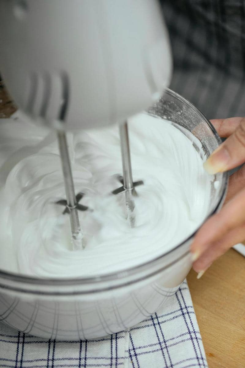 Tangan memegang mangkuk kaca berisi adonan putih yang sedang dikocok dengan mixer listrik untuk membuat cheesecake.
