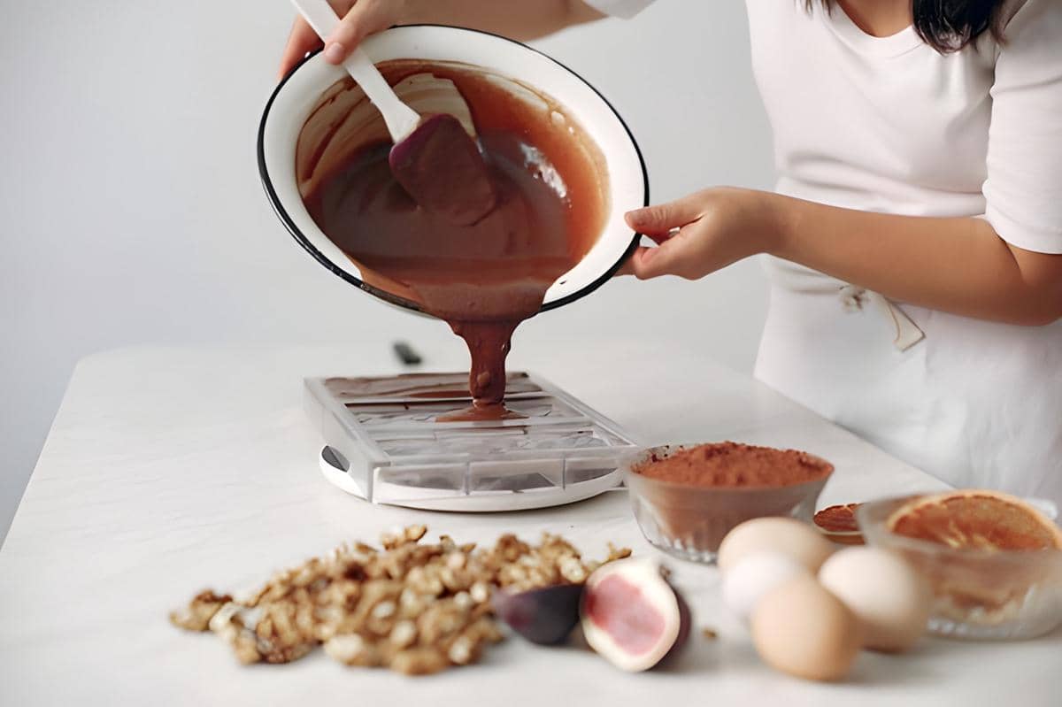 Seseorang menuang adonan cokelat ke dalam cetakan di meja dapur dengan bahan seperti telur, buah, dan bubuk kakao di sekitarnya.