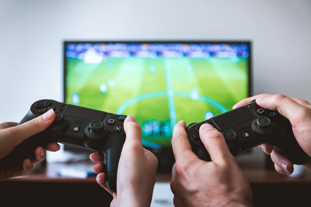 Dua orang memegang stik konsol PlayStation sambil bermain game sepak bola di layar televisi besar di ruang tamu.
