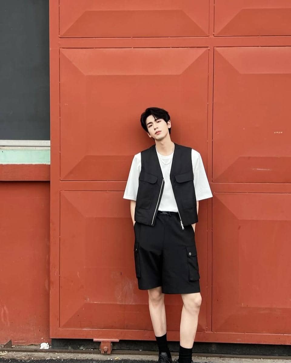Seorang pria berdiri bersandar di dinding merah mengenakan kaus putih, rompi hitam, celana pendek hitam, dan sepatu hitam.