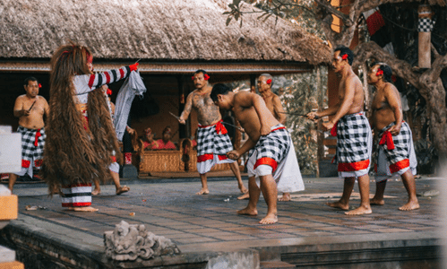 Beberapa penari pria mengenakan kain kotak hitam putih dan hiasan kepala merah sedang menampilkan pertunjukan tari tradisional di panggung terbuka.