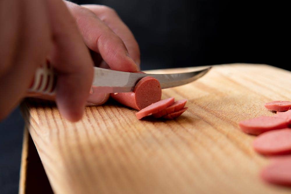 Tangan memotong sosis di atas talenan kayu menggunakan pisau dapur, dengan beberapa potongan sosis di sekitarnya.