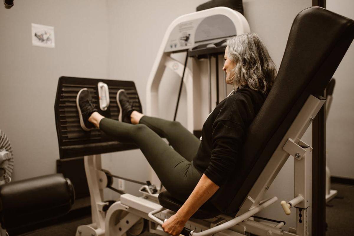 Seorang lansia perempuan sedang menggunakan alat leg press di gym.