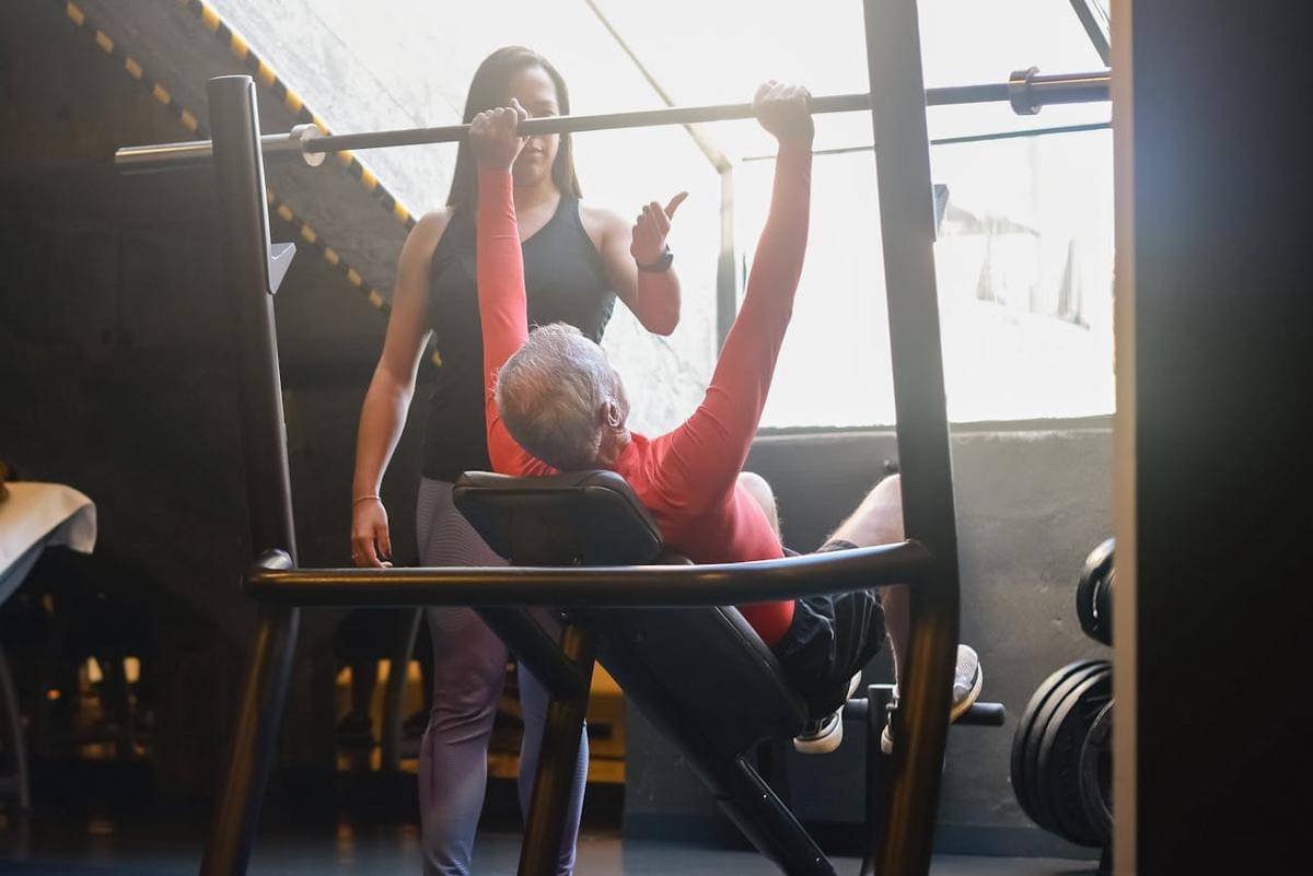 Seorang lansia laki-laki sedang berlatih bench press, ditemani seorang trainer perempuan.