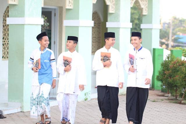 Empat santri berpakaian putih dan sarung berjalan bersama sambil membawa kitab di halaman pesantren dengan latar bangunan hijau.