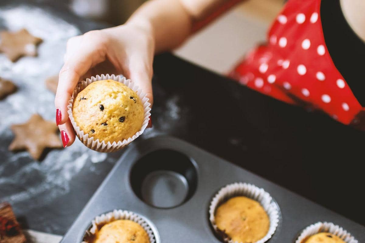Seseorang memegang muffin blueberry yang baru dipanggang di dapur dengan loyang muffin dan adonan kue di meja.