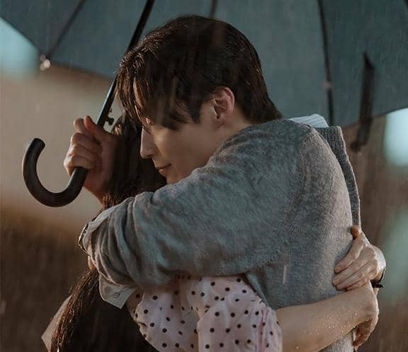 Dua orang berpelukan di bawah payung saat hujan dalam adegan drama A Hundred Memories, menciptakan suasana hangat dan emosional.