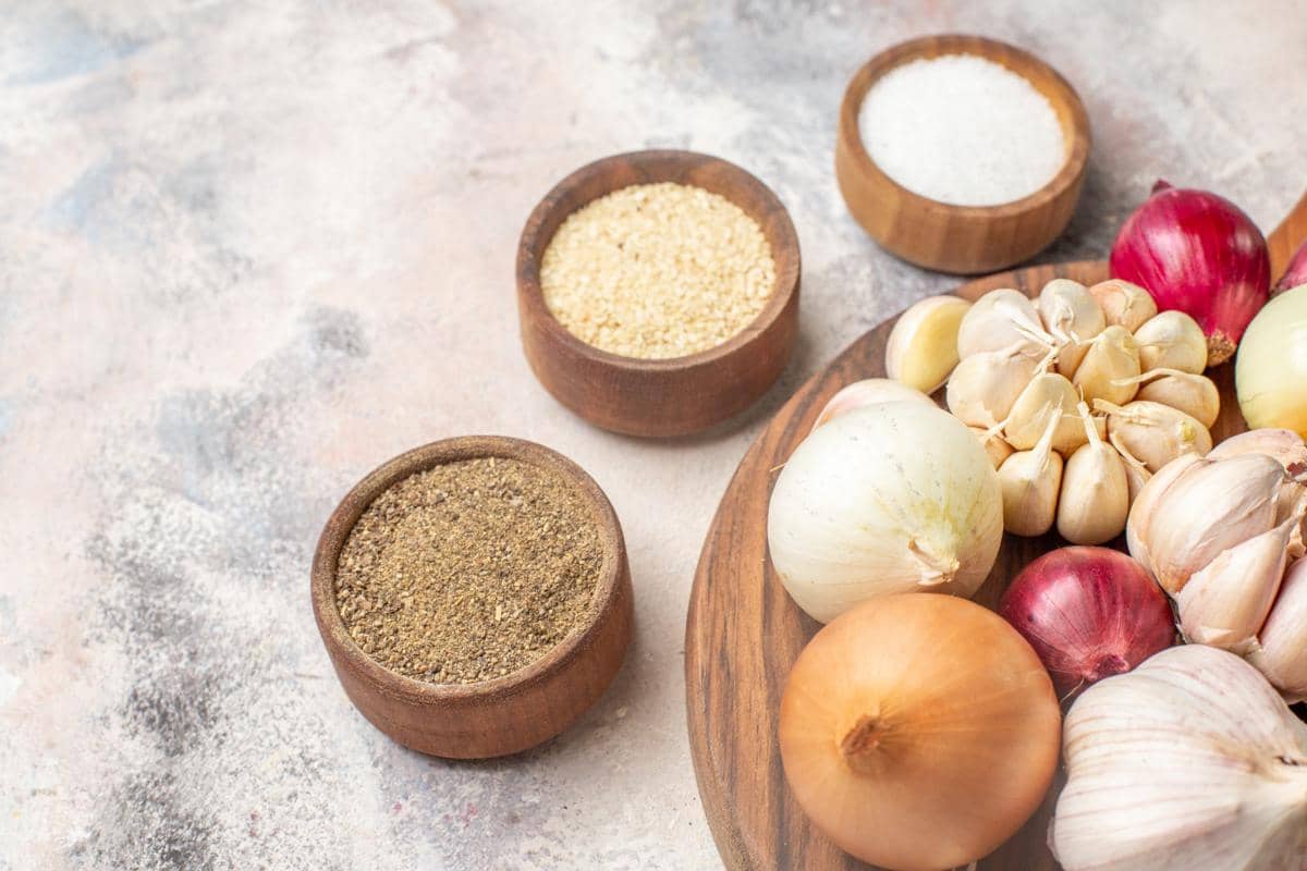 Bawang putih, bawang merah, dan bawang bombai di atas talenan kayu dengan mangkuk berisi garam, lada, dan bumbu dapur lainnya.