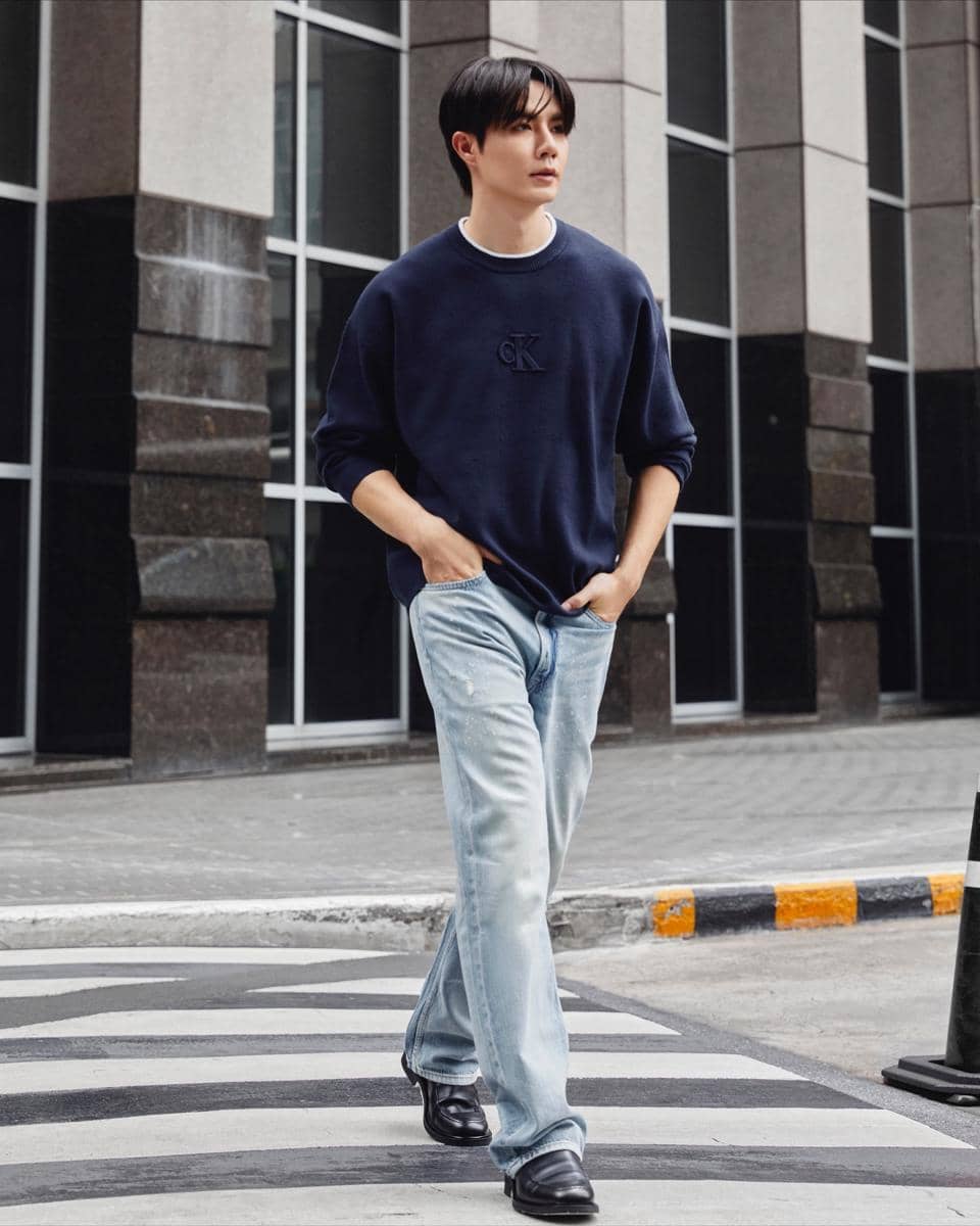 Pria mengenakan sweater biru tua dan celana jeans terang berjalan di trotoar kota dengan latar gedung kaca modern.