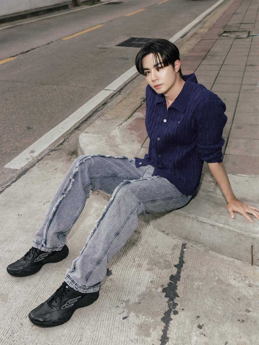 Seorang pria berpose santai di tepi trotoar mengenakan kemeja rajut ungu tua, celana jeans abu-abu, dan sepatu hitam.