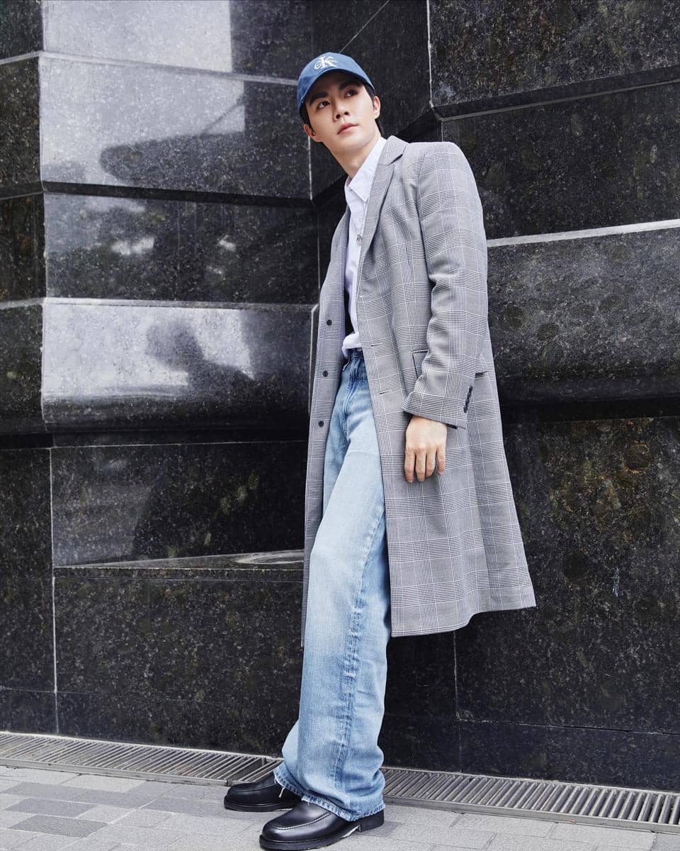 Pria mengenakan mantel abu-abu bermotif kotak, celana jeans longgar, kemeja putih, dan topi biru berdiri di depan dinding marmer hitam.