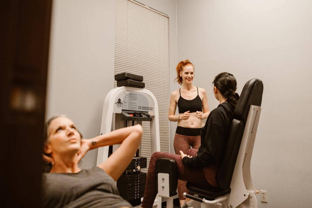 Seorang perempuan akan menggunakan mesin hip abduction di gym, dipandu oleh seorang trainer.
