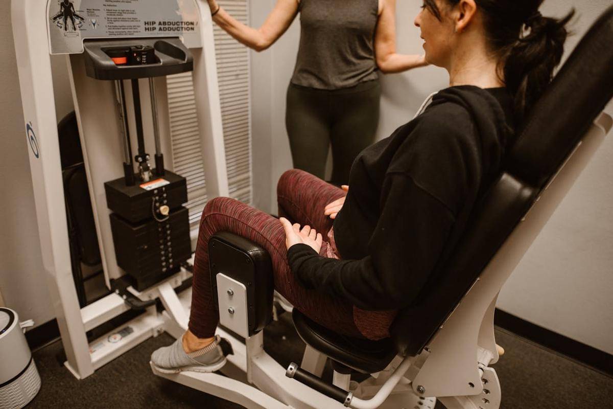 Seorang perempuan menggunakan mesin hip abduction di gym, didampingi dengan seorang trainer.