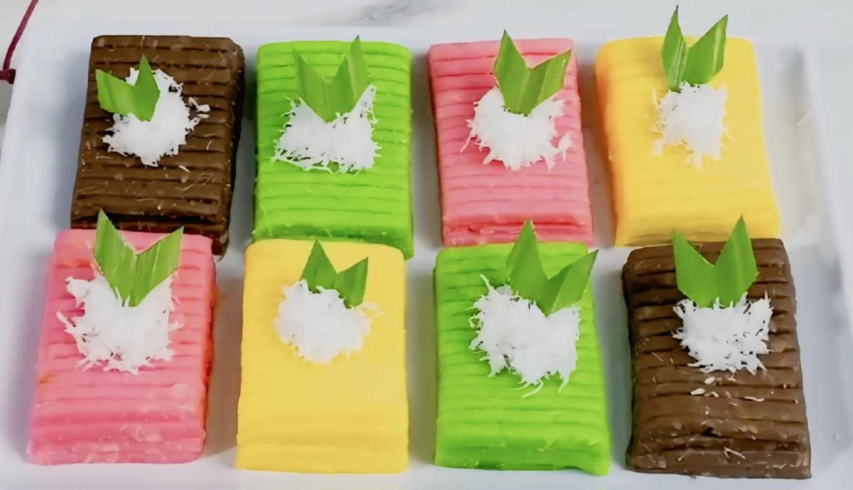 Delapan potong getuk lindri warna-warni disusun rapi di atas piring, masing-masing dihiasi parutan kelapa dan daun pandan di atasnya.