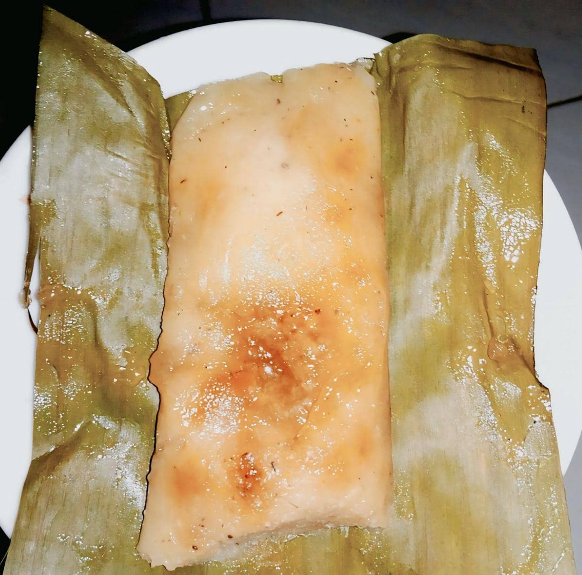 Lemet singkong berwarna cokelat muda dibungkus daun pisang yang terbuka di atas piring putih, tampak tekstur lembut dan mengilap.