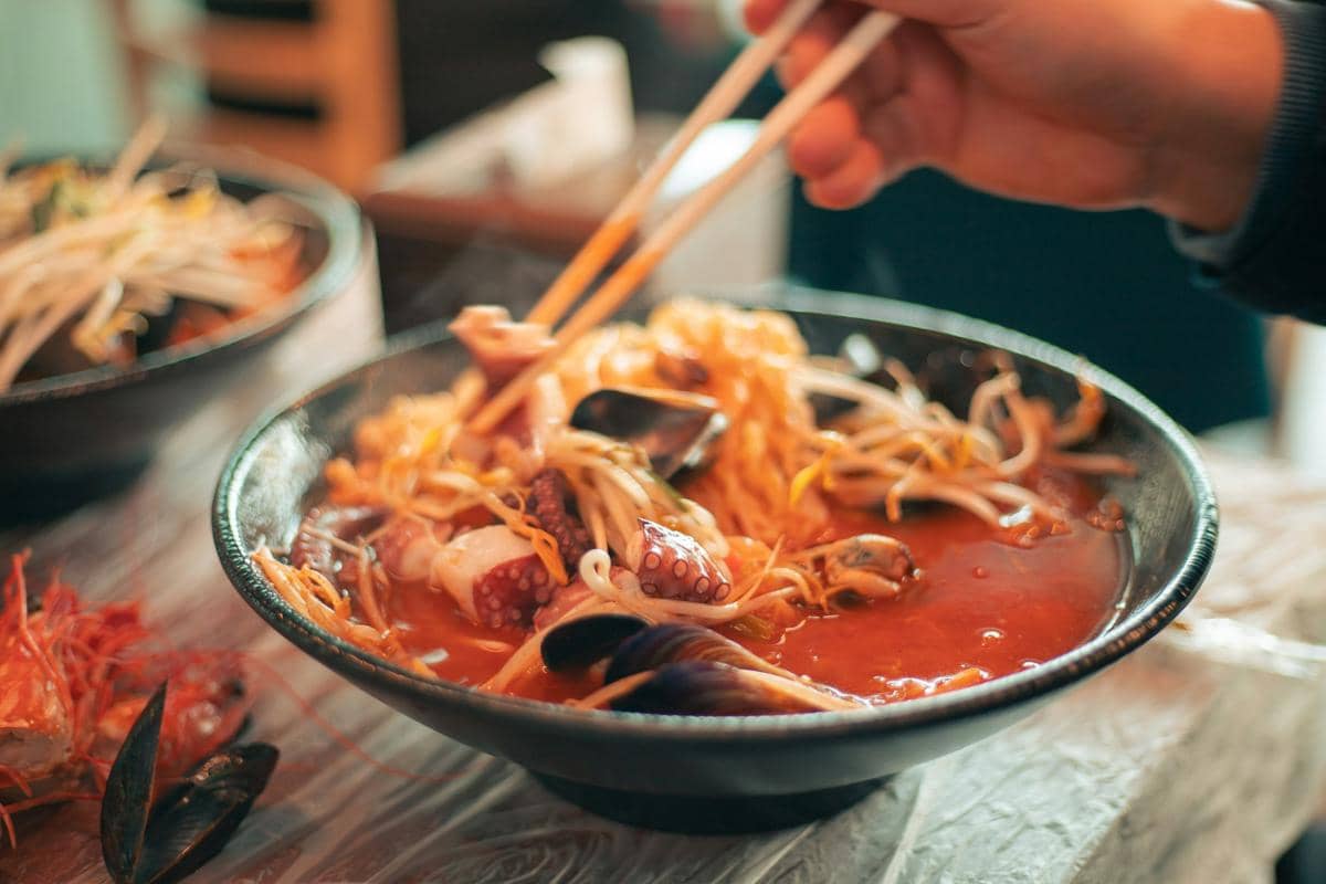 Semangkuk ramen dengan kuah merah berisi seafood seperti cumi, kerang, dan tauge, dipegang dengan sumpit di atas meja makan.