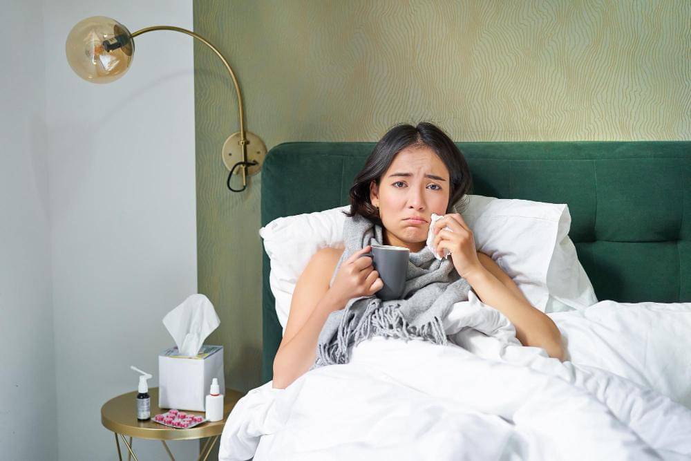 Seorang perempuan beristirahat di tempat tidur sambil meminum minuman hangat.