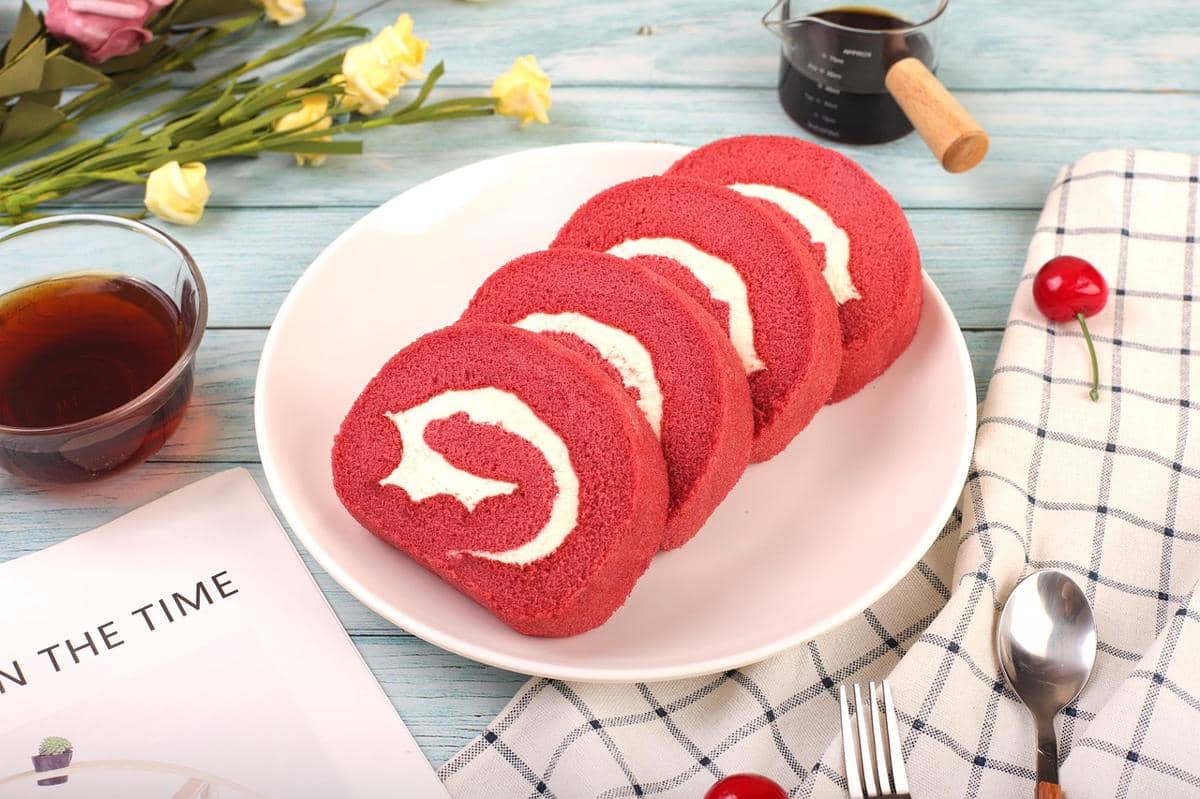 Empat potong kue gulung red velvet dengan isian krim putih tersaji di piring putih di atas meja kayu biru dengan minuman dan hiasan bunga.