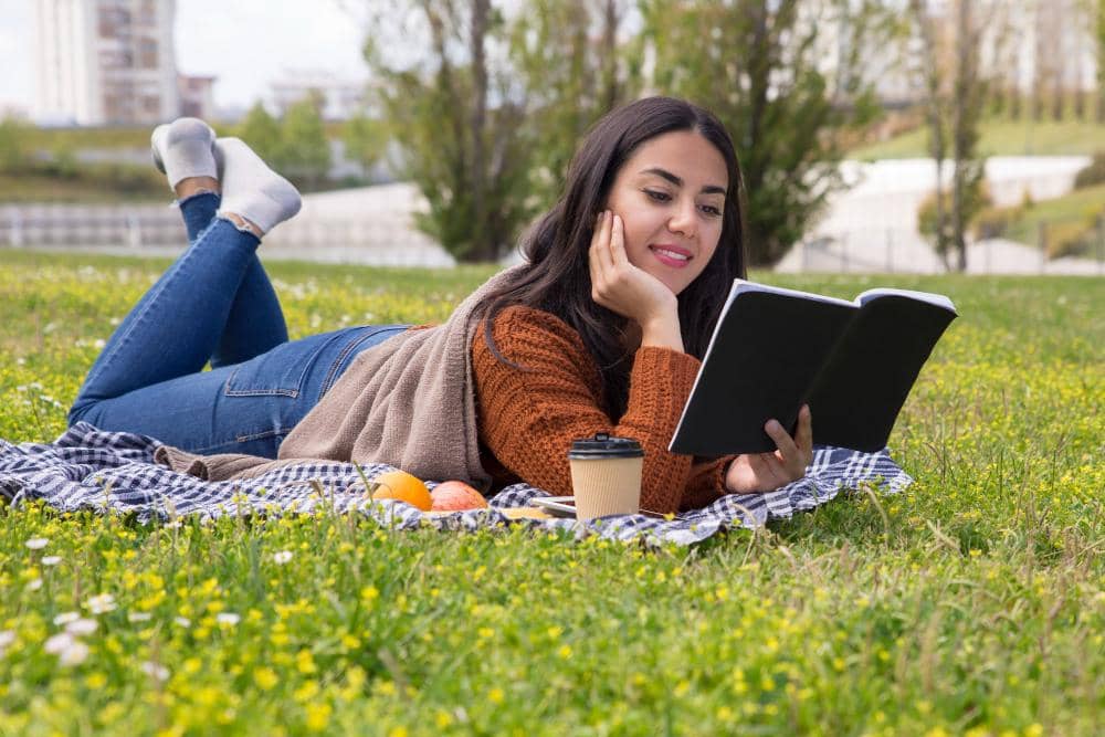 Seorang perempuan berbaring di atas selimut di taman sambil membaca buku, dengan kopi dan buah di sampingnya pada hari cerah.