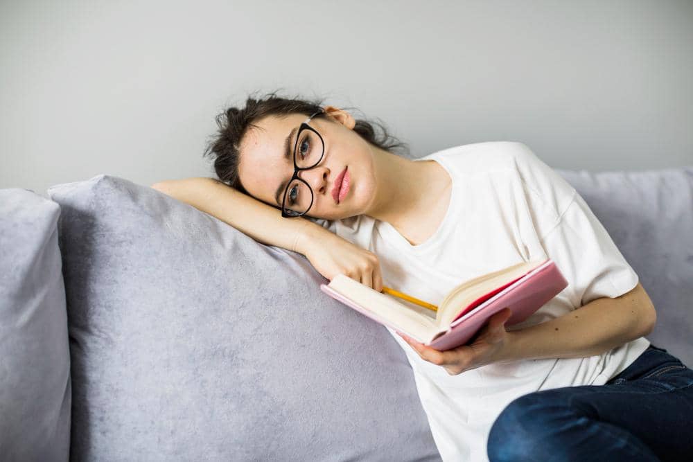 Perempuan berkacamata duduk di sofa dengan ekspresi jenuh sambil memegang buku catatan berwarna merah muda dan pensil di tangannya.