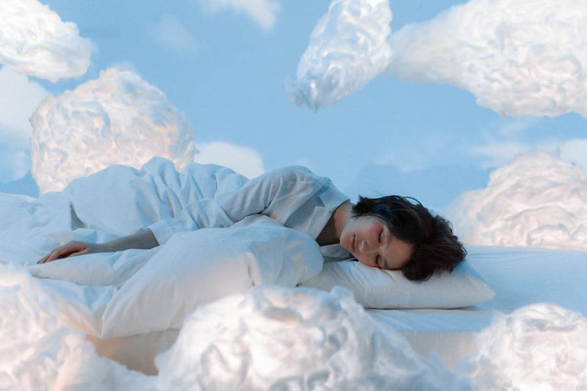 Seorang perempuan tidur nyenyak di atas ranjang dengan selimut, menggambarkan suasana bermimpi pada malam hari.