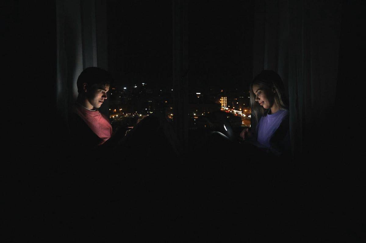Pasangan duduk di dekat jendela kamar tidur sambil melihat layar gadget, dengan latar belakang cahaya kota.