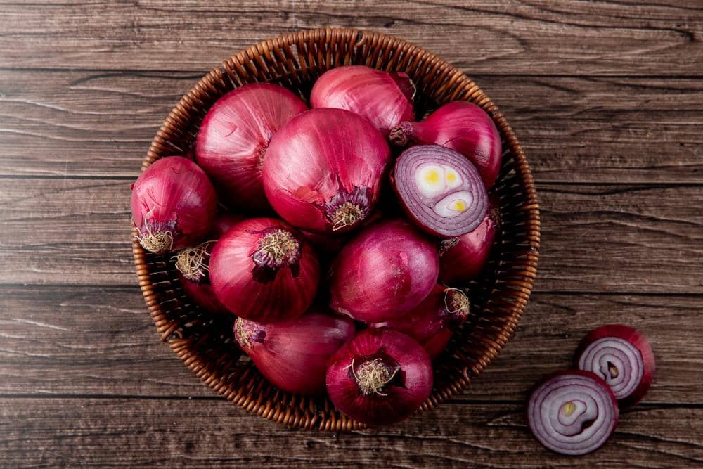 Beberapa bawang merah segar berwarna ungu kemerahan tersusun dalam keranjang anyaman di atas meja kayu cokelat.