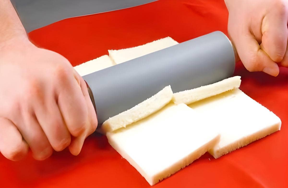 Tangan menggiling beberapa lembar roti tawar menggunakan rolling pin di atas alas berwarna merah.