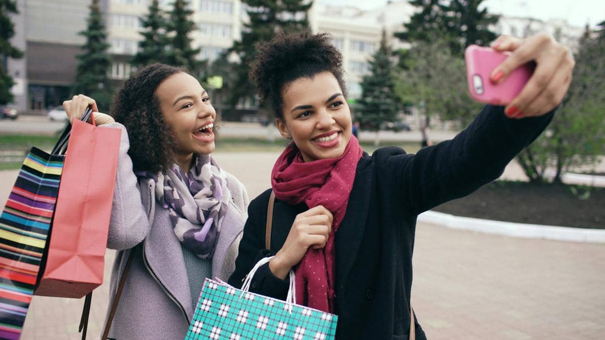 dua orang remaja sedang memotret diri mereka dengan pose memamerkan kantung belanja.