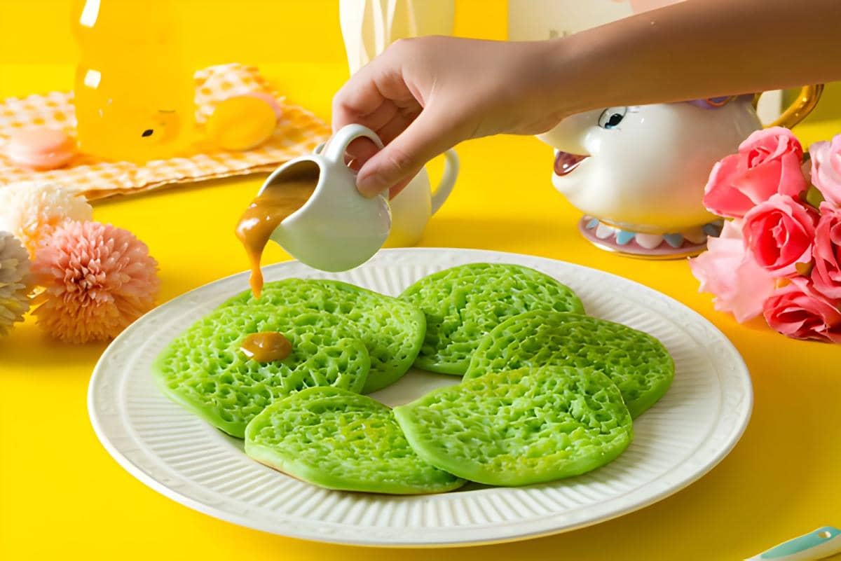 Serabi pandan hijau disiram kuah santan kental berwarna cokelat di atas piring putih dengan latar meja kuning cerah.