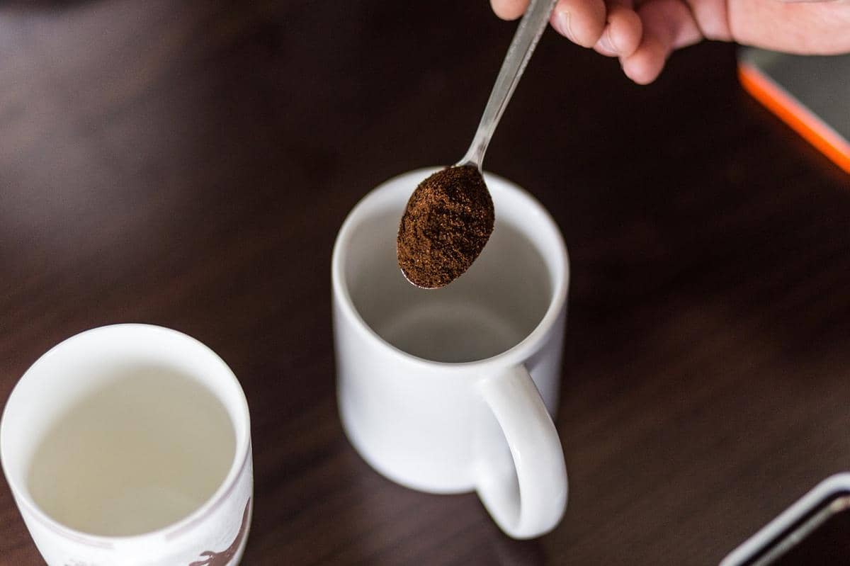 Seseorang menuangkan bubuk kopi ke dalam cangkir putih menggunakan sendok di atas meja kayu gelap.