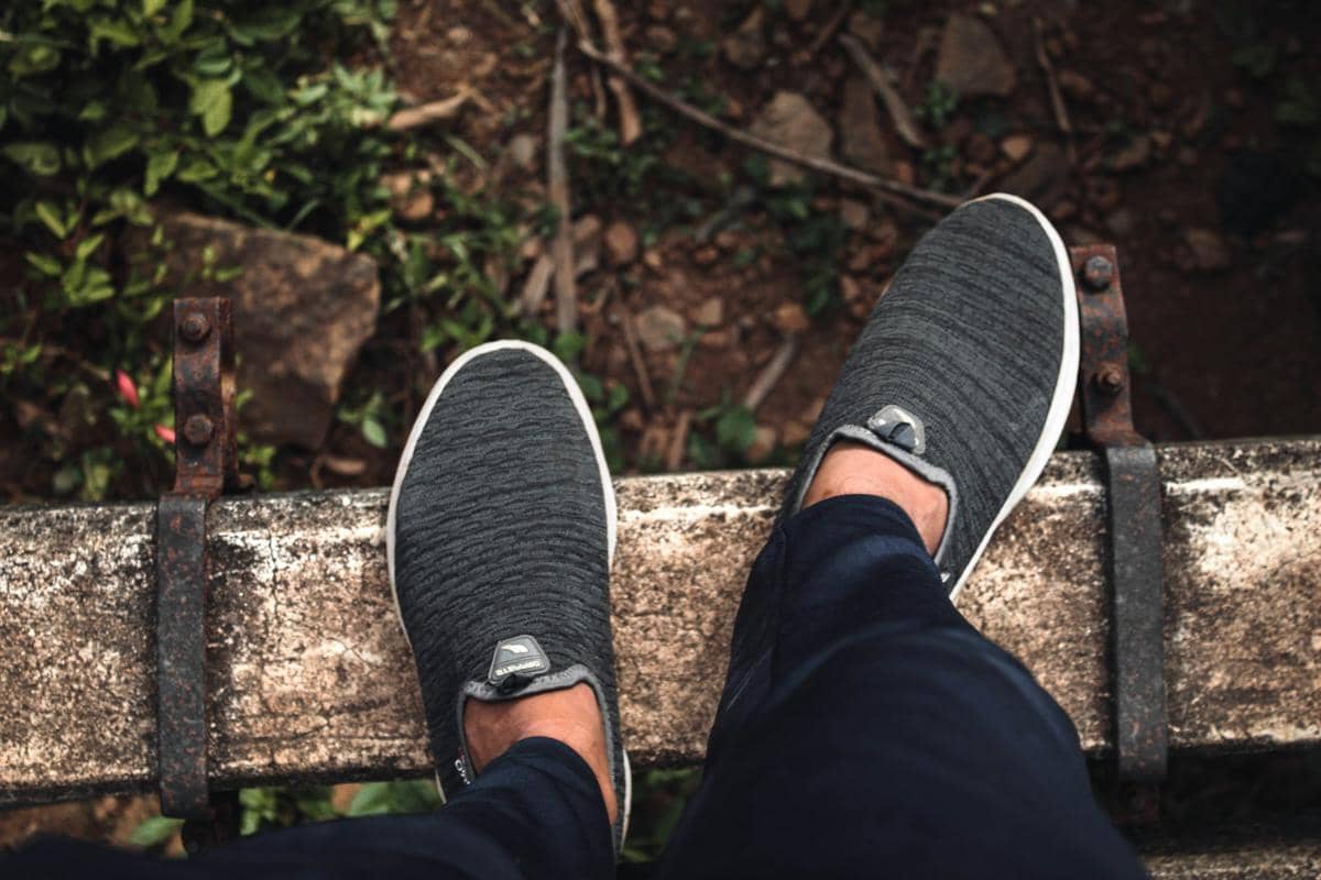 Sepasang kaki mengenakan sepatu slip-on berwarna abu-abu dengan sol putih, berdiri di atas rel besi tua di area outdoor.
