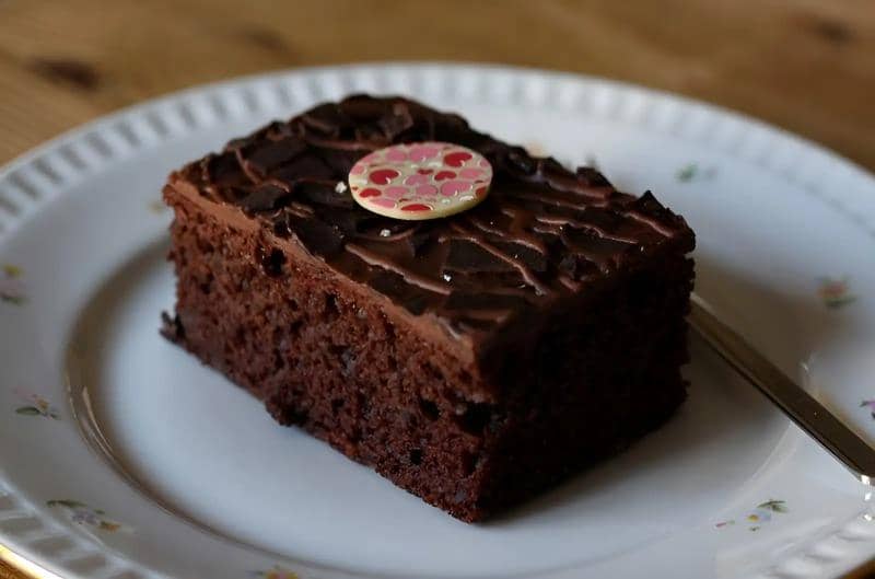 Sepotong brownies kukus cokelat dengan topping cokelat dan hiasan bulat di atasnya disajikan di atas piring putih bermotif bunga.