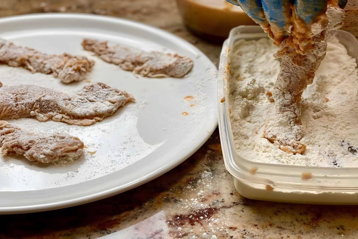 Potongan daging dilapisi tepung di atas piring dan wadah plastik di meja dapur, siap untuk digoreng atau dimasak lebih lanjut.