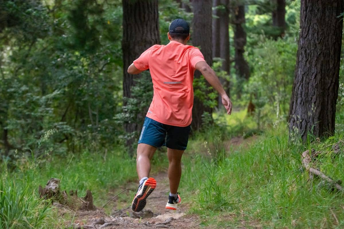 Seorang pria berlari di jalur hutan mengenakan sepatu trail running, kaus oranye, dan celana pendek biru gelap di tengah pepohonan hijau.
