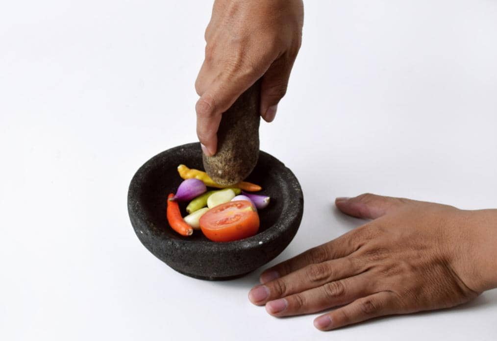 Tangan sedang mengulek bumbu dengan cobek batu berisi cabai, bawang, dan tomat di atas permukaan putih.