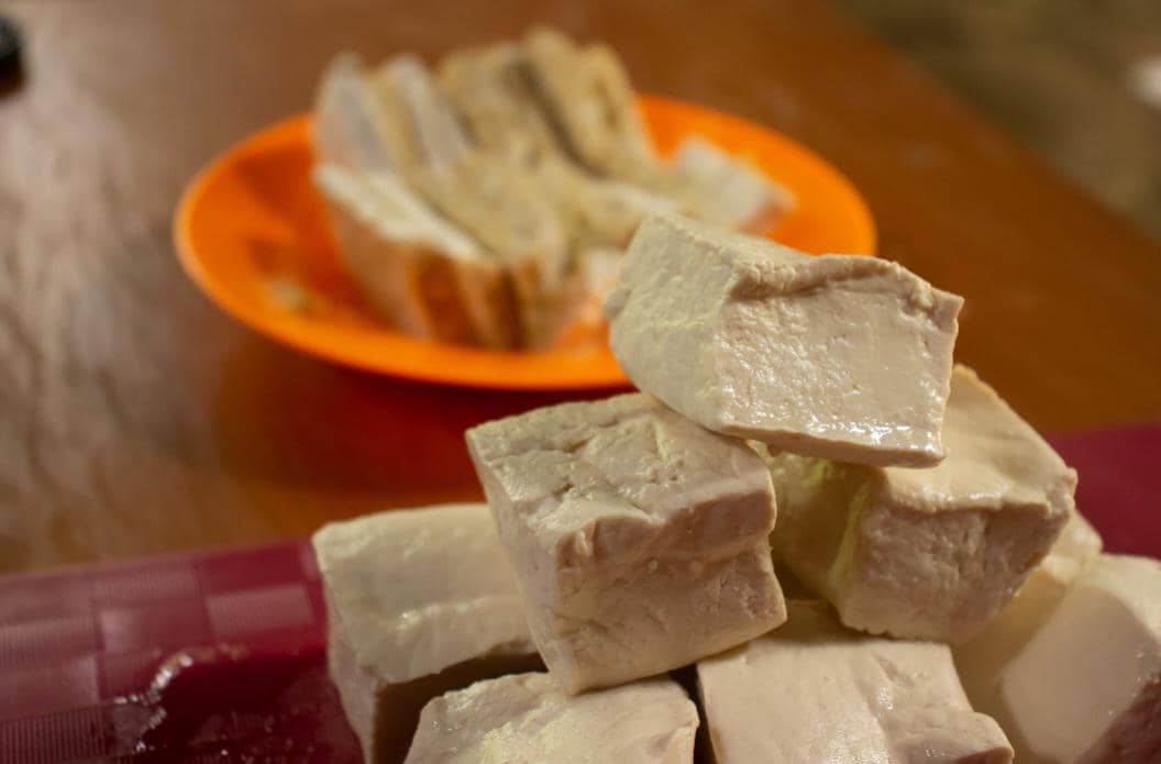 Potongan tahu putih segar disusun di atas talenan dengan latar belakang piring oranye berisi tahu goreng di meja kayu.