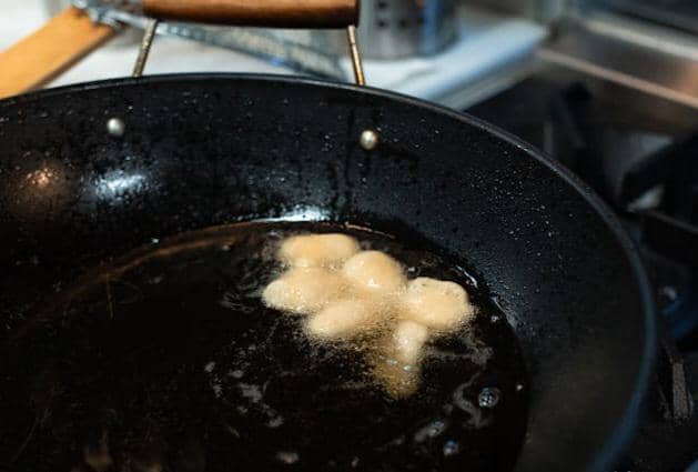 Beberapa potongan adonan digoreng dalam minyak panas di wajan hitam besar di atas kompor dapur modern.