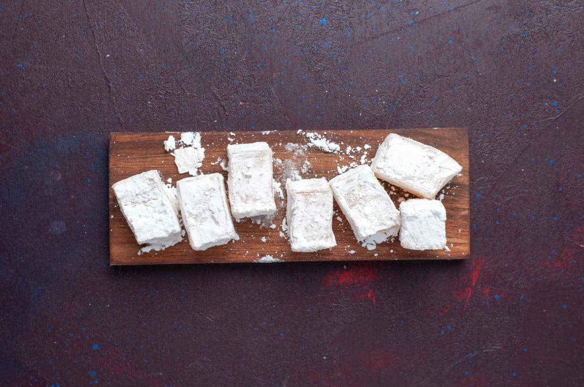Potongan tahu yang sudah dibaluri tepung terigu tersusun di atas talenan kayu dengan latar permukaan gelap bertekstur.