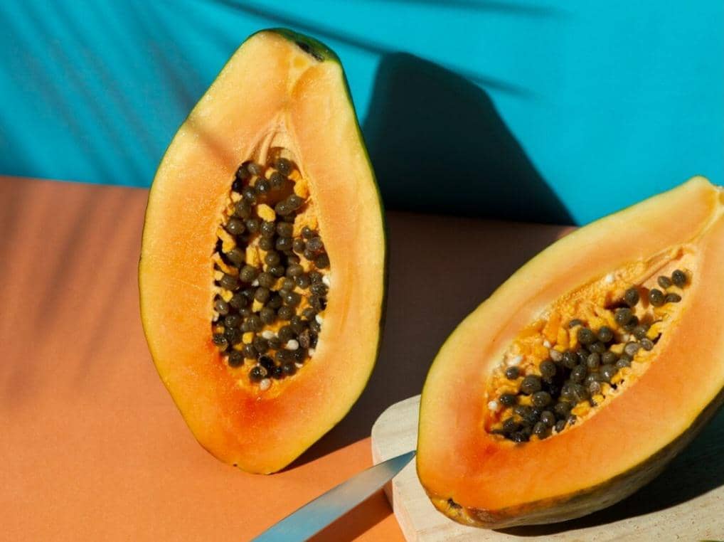 Dua potong pepaya muda berwarna oranye dengan biji hitam di atas permukaan berwarna oranye dan latar belakang biru cerah.