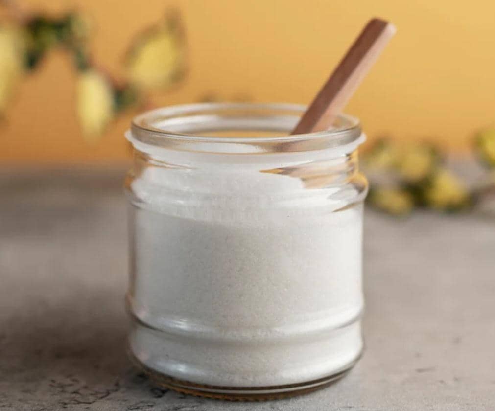 Stoples kaca berisi soda kue putih dengan sendok kayu kecil di dalamnya, diletakkan di atas permukaan abu-abu dengan latar oranye lembut.
