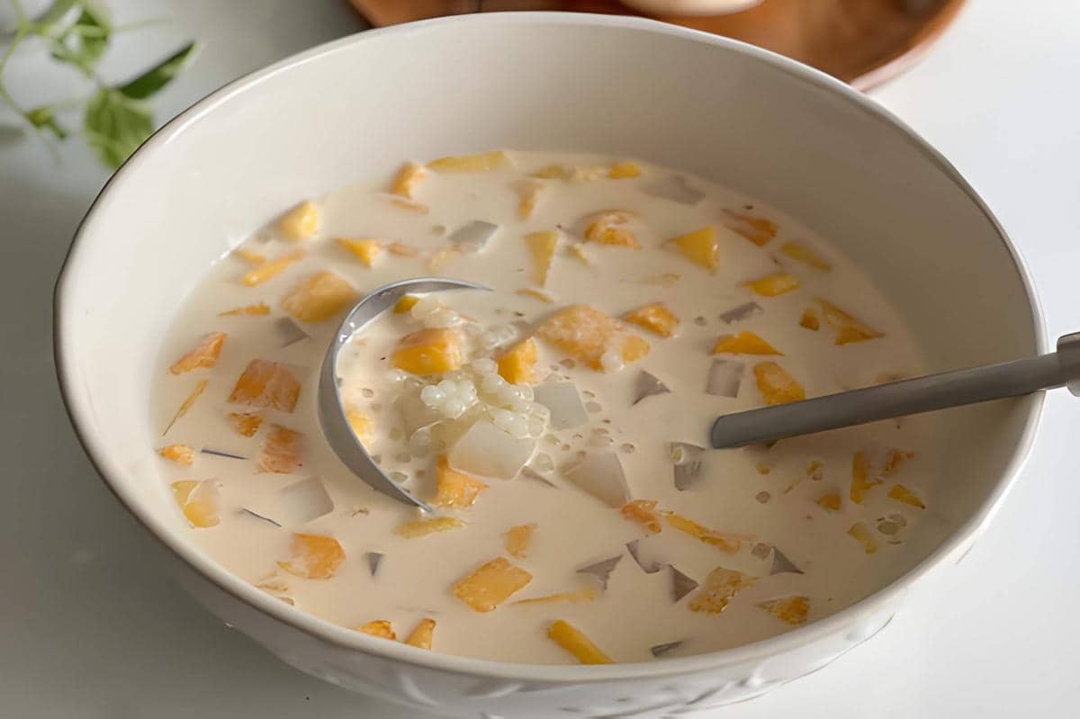 Semangkuk mango sago berisi potongan mangga, mutiara sagu, dan santan kental dengan sendok logam di dalamnya.