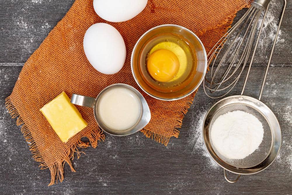 Ilustrasi bahan-bahan untuk membuat kue, termasuk tepung, telur, gula, dan peralatan dapur di atas meja.