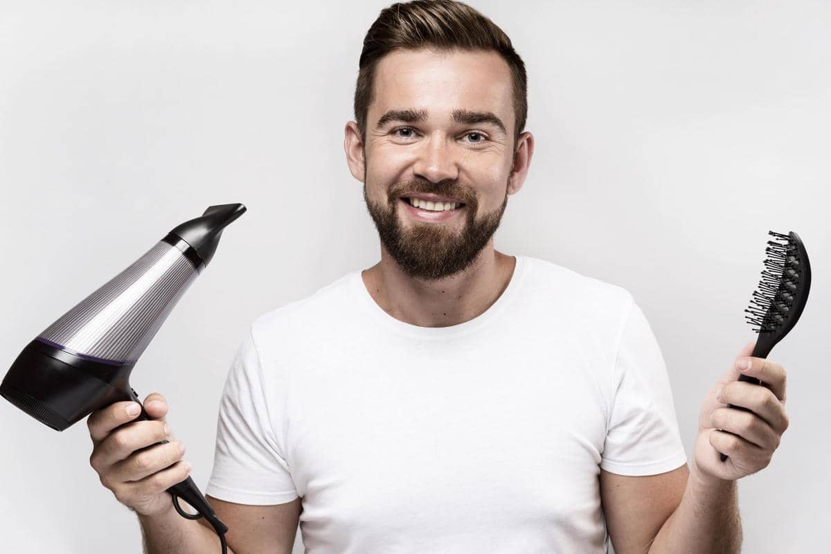 Pria berambut pendek dan berjanggut tersenyum sambil memegang pengering rambut di satu tangan dan sisir di tangan lainnya.