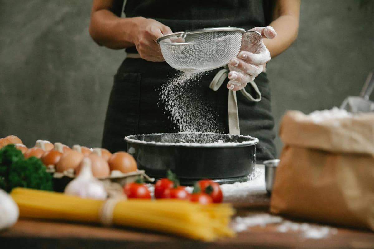 Seseorang mengenakan celemek sedang mengayak gula halus ke dalam loyang di dapur dengan bahan masakan seperti telur dan tomat di meja.